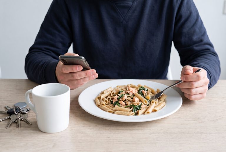 Sporter in donkerblauwe sweater houdt zijn telefoon vast tijdens het eten van penne met zalm en spinazie aan een houten keukentafel